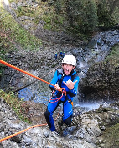 Junge Person beim Canyoning-Abenteuer in einer Naturwasserfalllandschaft, trägt Schutzhelm und Wetsuit. | © Rocksolid