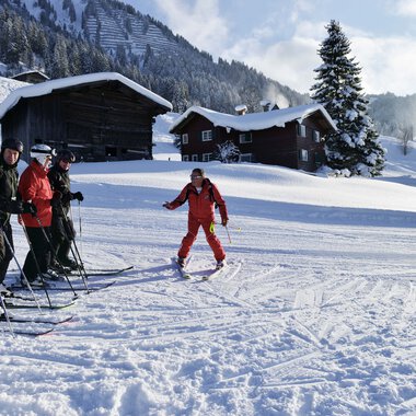 Skisports instructor and participants during a ski lesson in snowy mountains, instructor demonstrating a technique with background of cabins and snow-covered trees. | © Andy Herr