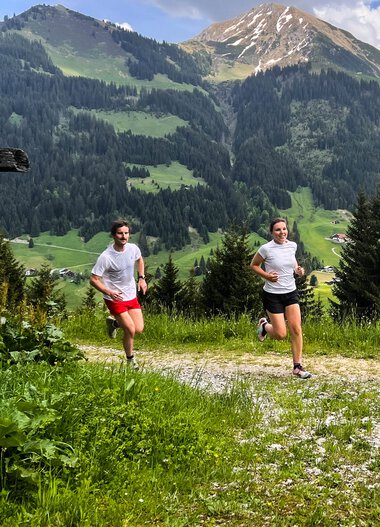Two people running on a trail in the mountains with green meadows and high peaks in the background. | © Kleinwalsertal Tourismus eGen 