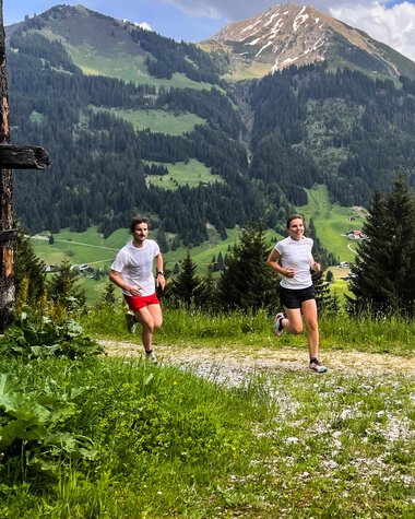Zwei Menschen laufen auf einem Waldweg in den Bergen mit grünen Wiesen und hohen Gipfeln im Hintergrund. | © Kleinwalsertal Tourismus eGen 