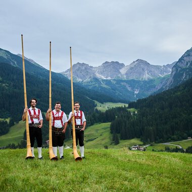 Drei Männer in Trachten stehen in einer grünen Wiese mit Alphörnern, umgeben von Bergen und einer bewaldeten Talregion. | © Kleinwalsertal Tourismus eGen • Fotograf: Oliver Farys